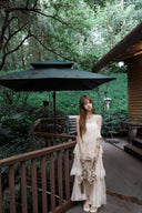 Woman in a white dress standing in a garden setting with trees and a building in the background.