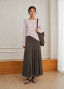 A woman stands indoors wearing a light pink long-sleeve top, a VOGSION Celine High Waist Silk-Flow Maxi Skirt, and black sandals, while carrying a dark brown shoulder bag.