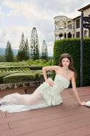 Woman in a Vogsion Pistachio Green Strapless Corset Ruffle Peplum Lace Prom Dress 2026 lying on a wooden deck with a scenic background