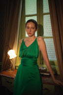 Woman in a green dress standing in a room with curtains and a lamp.