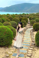 Woman in Vogsion "Lucienne" Ethereal Tulle & Contrast Lace-Inset Midi Dress standing on a stone path through a neatly trimmed hedge maze with mountains in the background.