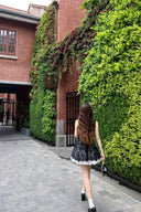 Woman in Vogsion Shush Trong Renaissance Stiff Wool-Lace Coquette Dress walking along a path lined with greenery and a brick building.