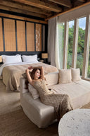 Woman wears Vogsion Champagne Gold Cross-Strap Backless Mermaid Maxi Dress relaxing on a beige sofa in a stylish bedroom with large windows and wooden ceiling.