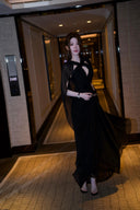Vogsion Open Back Ribbon Maxi Dress – Elegant black pleated chiffon halter neck evening gown with dramatic sheer ribbon cape sleeves, open back cutouts, and flowing maxi skirt, worn by real Vogsion customer (not professional model) in luxury setting with global fast shipping