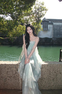 Woman in a Vogsion Seraphina Tiered Chantilly Lace Maxi Dress standing by a river with trees and buildings in the background