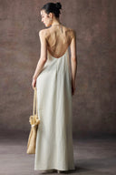 Woman in Vogsion Architectural Column Gown in sand with an open back, holding a woven bag against a brown background.