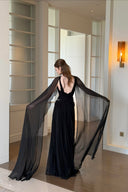 Vogsion Open Back Ribbon Maxi Dress – Elegant black pleated chiffon halter neck evening gown with dramatic sheer ribbon cape sleeves, open back cutouts, and flowing maxi skirt, worn by real Vogsion customer (not professional model) in luxury setting with global fast shipping