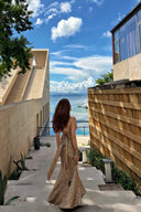 Woman in a Vogsion Champagne Gold Cross-Strap Backless Mermaid Maxi Dress walking along a path by a building with a view of the ocean.