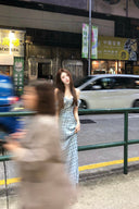 Woman in a blue polka dot dress standing on a street with blurred pedestrians and vehicles in the background.