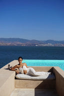 Person lounging on a sunbed by an infinity pool with a scenic view of mountains and sea in Vogsion ivory strapless cut out ruched maxi dress