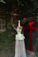 Woman in a Vogsion Pistachio Green Strapless Corset Ruffle Peplum Lace Prom Dress 2026 standing in front of a door with a red ribbon, surrounded by greenery.