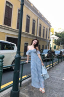 Woman in a blue polka dot dress standing on a street with buildings and cars in the background.