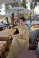 Woman in a Vogsion Light Pomelo Olive Mesh Knit Draped Cape Pleated Maxi Dress sitting at an outdoor cafe with palm trees in the background