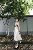 Woman in Vogsion "Lucienne" Ethereal Tulle & Contrast Lace-Inset Midi Dress with black floral details standing outdoors near a tree and wall.