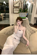 Woman in Vogsion Élodie Bow-Tiered Ivory Lace Maxi Dress sitting on a beige sofa in an elegant room.