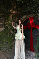 Woman in a Vogsion Pistachio Green Strapless Corset Ruffle Peplum Lace Prom Dress 2026 standing in front of a door with a large red bow, surrounded by greenery.