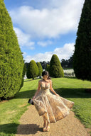 Woman wearing the VOGSION Luna Twist Midi Dress in Cream Perla Paisley silk-linen fabric, featuring a voluminous flare skirt and waist cut-out in a lush garden setting