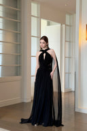 Vogsion Open Back Ribbon Maxi Dress – Elegant black pleated chiffon halter neck evening gown with dramatic sheer ribbon cape sleeves, open back cutouts, and flowing maxi skirt, worn by real Vogsion customer (not professional model) in luxury setting with global fast shipping