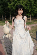 Woman in a Vogsion Odette ethereal 3D floral watercolor mesh maxi dress holding a beige bag outdoors with greenery in the background