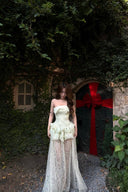 Woman in a Vogsion Pistachio Green Strapless Corset Ruffle Peplum Lace Prom Dress 2026 standing in front of a stone archway with a large red bow.