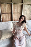 Woman in a Vogsion Sunset Aurelia Lace-Trimmed Liquid Satin Gown – Champagne Pink Resort Maxi Slip Dress sitting on a white couch with wooden paneling in the background