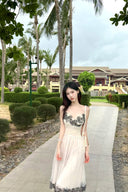 Woman in a Vogsion "Lucienne" Ethereal Tulle & Contrast Lace-Inset Midi Dress with black floral details standing on a path with palm trees and buildings in the background.