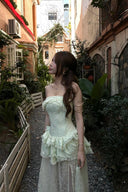 Woman in a Vogsion Pistachio Green Strapless Corset Ruffle Peplum Lace Prom Dress 2026 standing in an alleyway with plants and buildings.