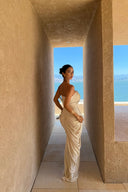Woman in a Vogsion ivory strapless cut out ruched maxi dress standing in a sunlit architectural setting with ocean view