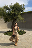Woman wearing the VOGSION Luna Twist Midi Dress in Cream Perla Paisley silk-linen fabric, featuring a voluminous flare skirt and waist cut-out in a lush garden setting