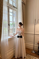 Woman in a Vogsion Élodie Lace Corset Bow Satin Maxi Dress with a black bow standing by a large window in an elegant room.