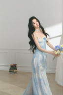 Woman in a light blue floral dress holding flowers in a minimalistic room.