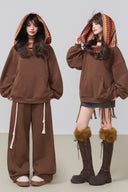 Two people wearing brown hoodies and pants with fringes on a plain background