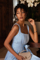 Model seated indoors wearing the Azure Linen Sky-Blue/Beige A-Line Backless Pearl Wide-Leg Jumpsuit with a square neckline, pearl back chain, wide-leg silhouette, waist tie and a neutral top-handle handbag.