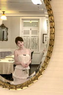 Woman in a Vogsion Élodie Bow-Tiered Ivory Lace Maxi Dress standing in front of a large decorative mirror in an elegant room.