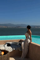 Woman in a Vogsion ivory strapless cut out ruched maxi dress standing by an infinity pool with a scenic view of mountains and water.