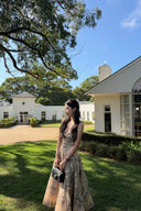 Woman wearing the VOGSION Luna Twist Midi Dress in Cream Perla Paisley silk-linen fabric, featuring a voluminous flare skirt and waist cut-out in a lush garden setting