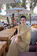 Woman in a Vogsion Light Pomelo Olive Mesh Knit Draped Cape Pleated Maxi Dress sitting outdoors with palm trees and tables in the background