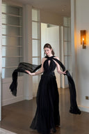 Vogsion Open Back Ribbon Maxi Dress – Elegant black pleated chiffon halter neck evening gown with dramatic sheer ribbon cape sleeves, open back cutouts, and flowing maxi skirt, worn by real Vogsion customer (not professional model) in luxury setting with global fast shipping