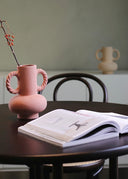 Pink vase on a table with an open book and another vase in the background.