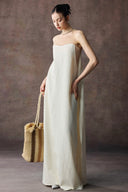 Woman in Vogsion Architectural Column Gown in sand holding a straw bag against a textured brown background