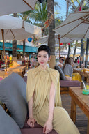 Woman in a Vogsion Light Pomelo Olive Mesh Knit Draped Cape Pleated Maxi Dress sitting on a bench with a tropical setting in the background
