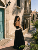 Back view of VOGSION Riviera Muse Deep V Halter Backless Maxi Dress featuring an open back, nautical ivory trim, flowing black maxi skirt and elegant European resort architecture.