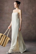 Woman in Vogsion Architectural Column Gown in sand holding a straw bag against a textured brown background