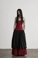 A model with long black hair and visible arm tattoos stands in a studio wearing a gothic-style dress with a red bodice, a voluminous black skirt, and a red ruffled underskirt.