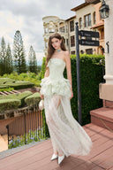 Woman in a Vogsion Pistachio Green Strapless Corset Ruffle Peplum Lace Prom Dress 2026 standing on a wooden deck with greenery and buildings in the background.