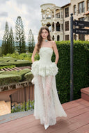 Woman in a Vogsion Pistachio Green Strapless Corset Ruffle Peplum Lace Prom Dress 2026 standing on a wooden deck with greenery and buildings in the background.