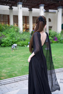 Vogsion Open Back Ribbon Maxi Dress – Elegant black pleated chiffon halter neck evening gown with dramatic sheer ribbon cape sleeves, open back cutouts, and flowing maxi skirt, worn by real Vogsion customer (not professional model) in luxury setting with global fast shipping
