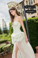 Woman in a Vogsion Pistachio Green Strapless Corset Ruffle Peplum Lace Prom Dress 2026 standing outdoors with greenery and buildings in the background