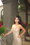 Woman in a Vogsion Champagne Gold Cross-Strap Backless Mermaid Maxi Dress standing outdoors with decorative columns and flowers in the background