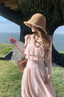 Woman in a pink Vogsion lace-trim ruffle maxi dress and straw hat standing by a tree with a scenic background